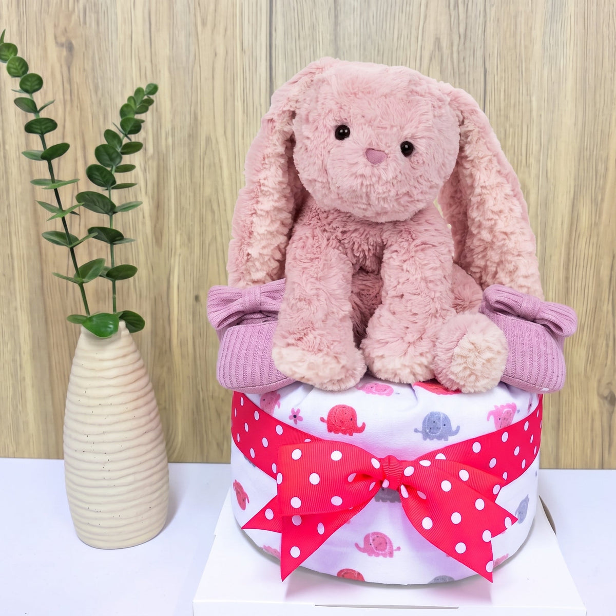 A pink plush bunny toy seated on top of a wrapped diaper cake with a red polka-dot ribbon and bow, styled on a white surface against a wood-look wall with a small vase of greenery beside it.
